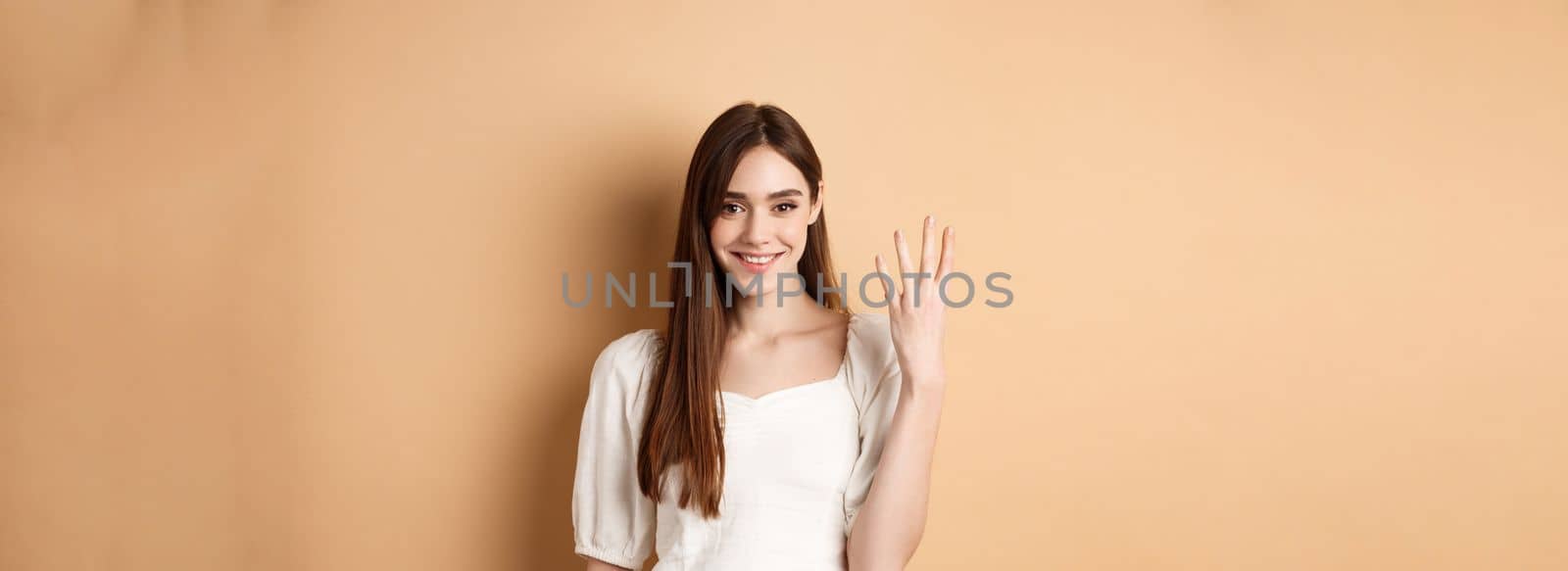 Attractive young woman show fingers number four, smiling and looking confident, standing on beige background.