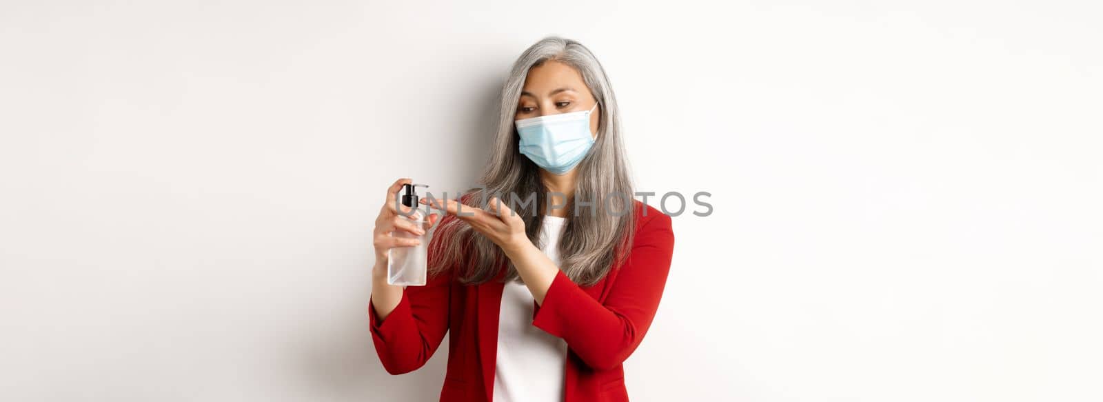 Covid, pandemic and business concept. Businesswoman in red blazer and face mask using hand sanitizer to clean and disinfect, standing over white background.