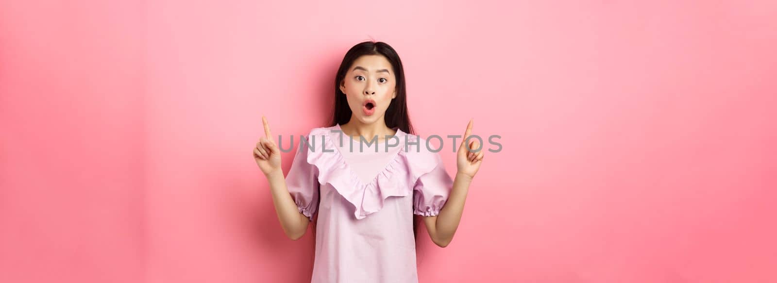 Excited asian teen girl pointing fingers up, saying wow and showing promo deal, standing in dress against pink background.