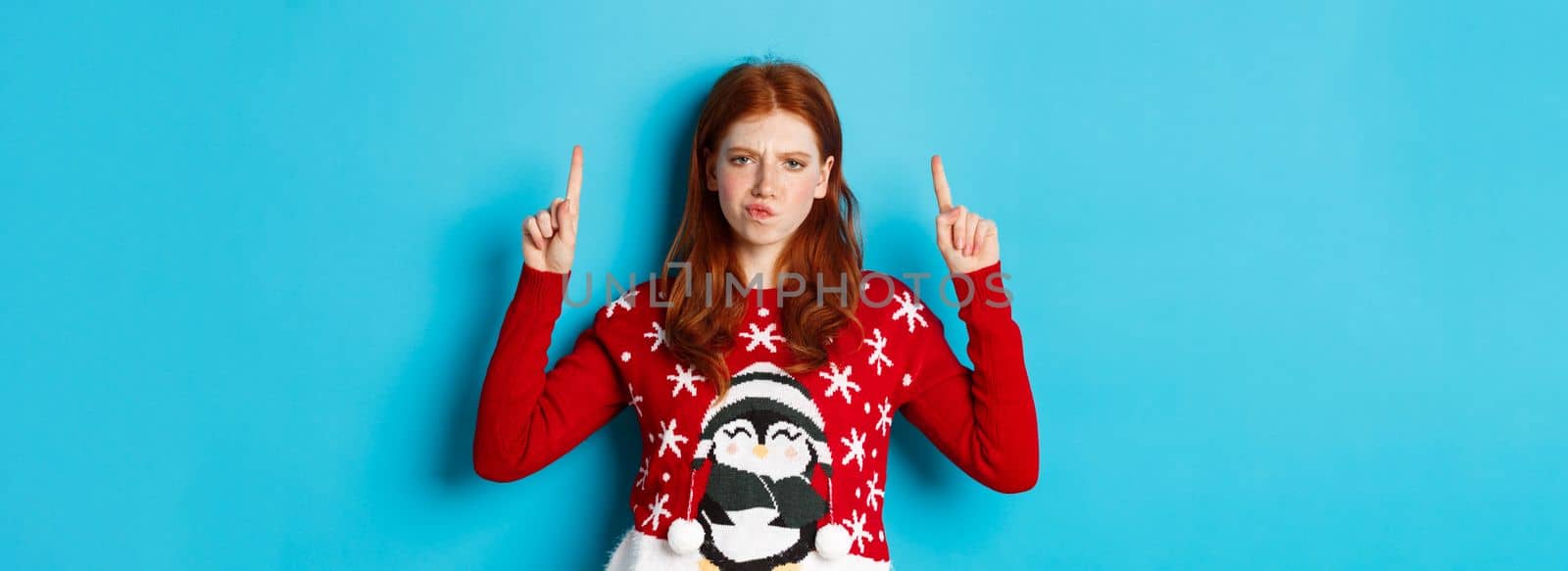 Winter holidays and celebration concept. Perplexed redhead girl in xmas sweater, frowning doubtful and pointing fingers up, showing advertisement, blue background.