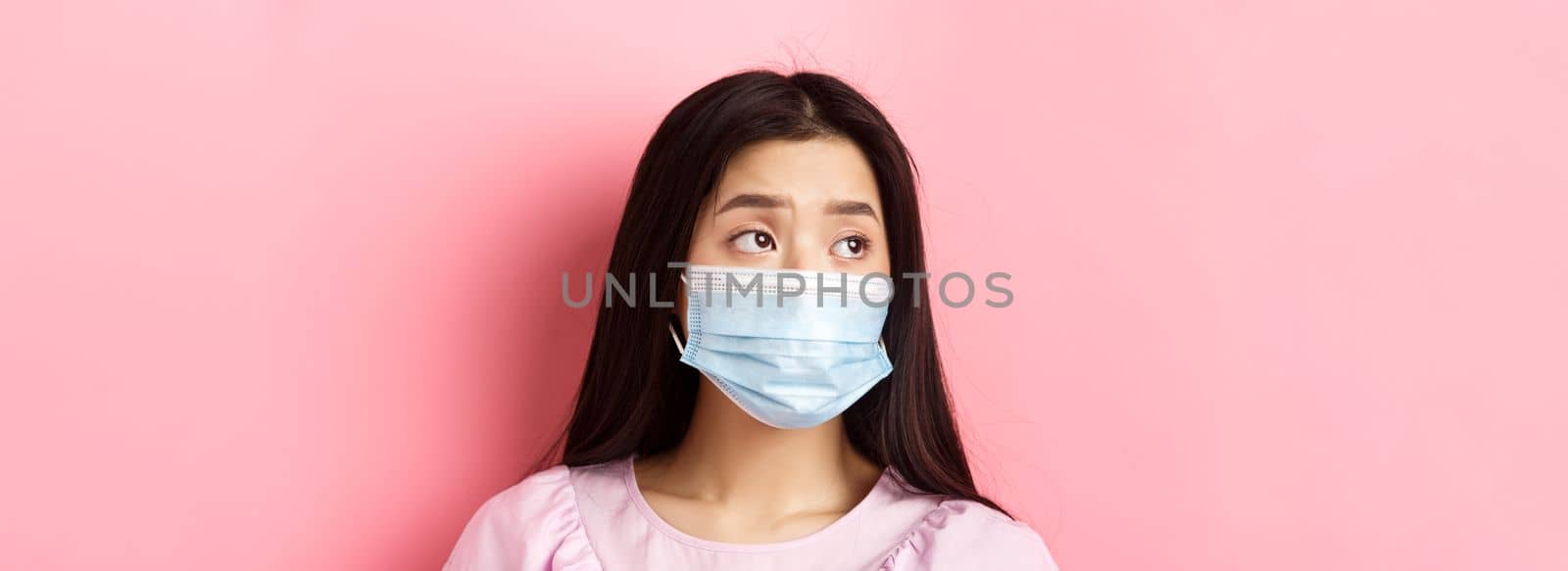 Covid-19, pandemic and quarantine concept. Close-up of sad asian girl in medical mask feeling lonely during coronavirus, standing against pink background by Benzoix