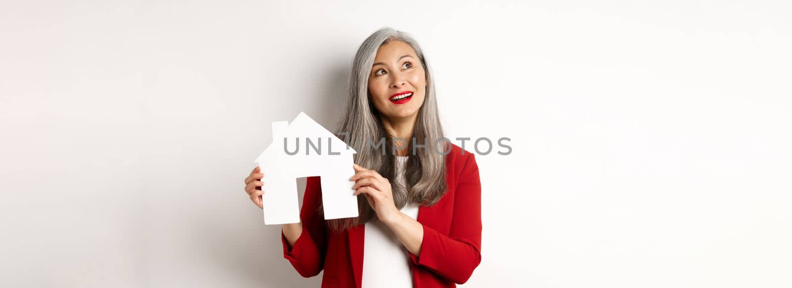 Dreamy senior woman thinking of buying property, showing paper house cutout and looking at upper left corner, standing over white background.