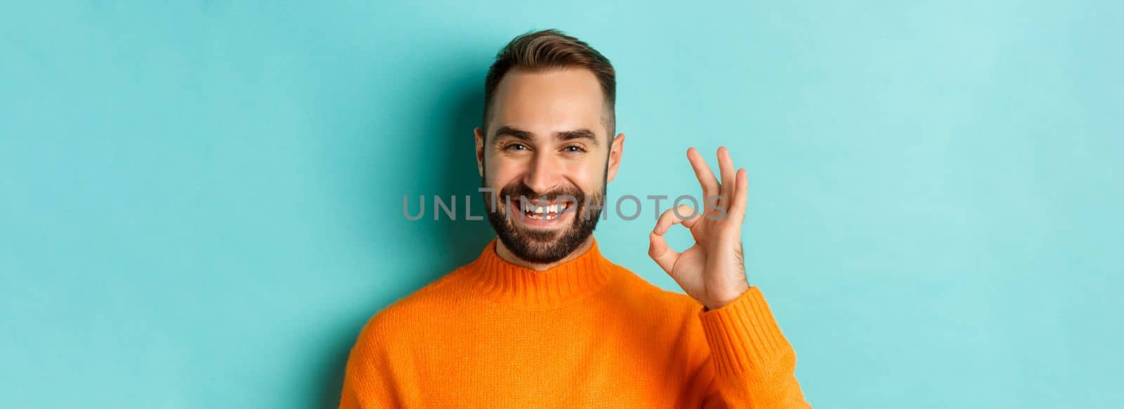 Close-up of assertive young man assuring everything ok, showing okay sign and smiling, yes or positive answer, standing over light blue background.