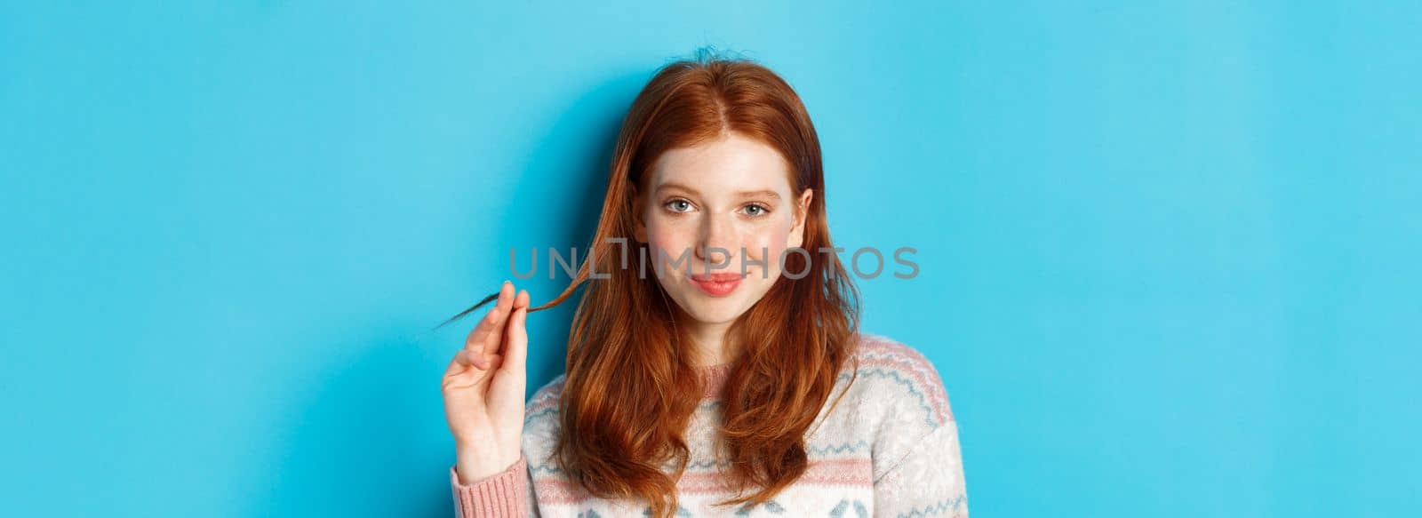 Close-up of confident and sassy redhead teen girl looking at camera pleased, playing with hair strand and smirking, standing over blue background.