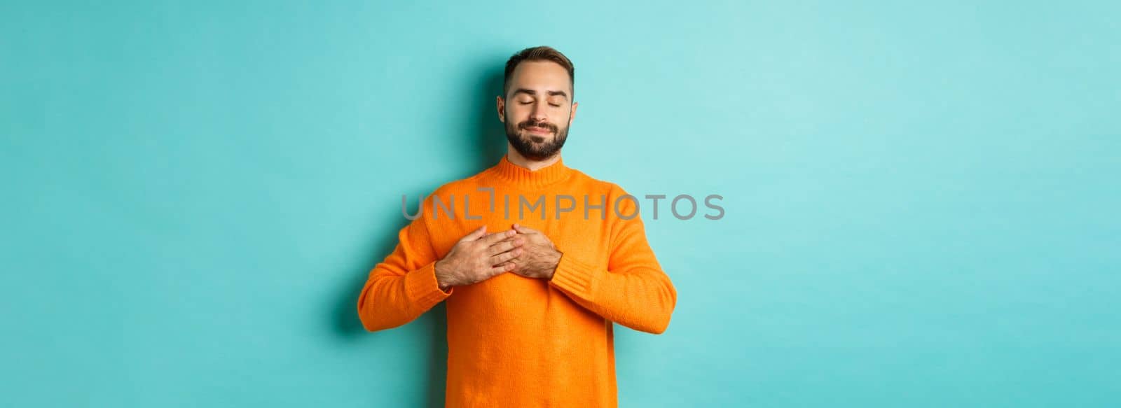 Handsome man feeling nostalgic, holding hands on heart and daydreaming, remember or imaging something, standing carefree against turquoise background.