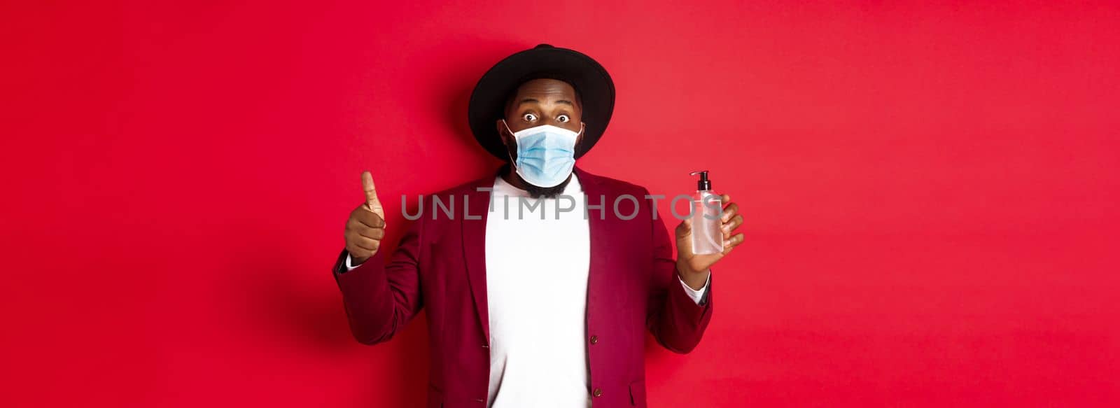 Covid-19, quarantine and holidays concept. Impressed black man in party outfit and medical mask, showing thumb up and hand sanitizer, standing over red background by Benzoix