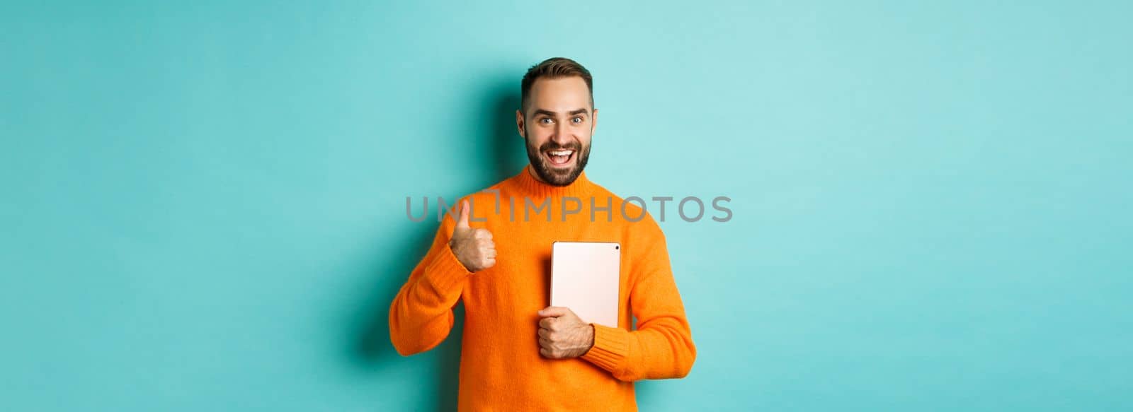 Work from home, technology concept. Handsome man holding laptop, showing thumb up, approve and like something, standing over turquoise background.