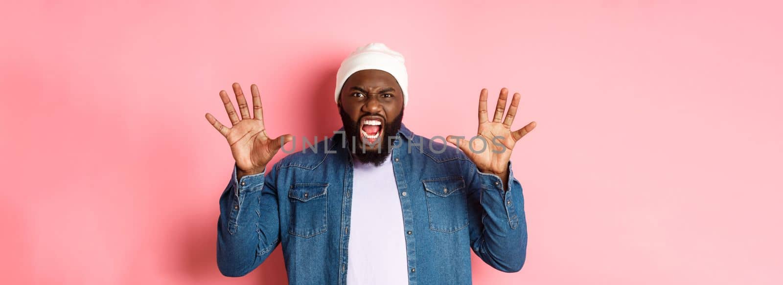 Angry african-american guy in beanie, scare you, roaring and screaming, showing hands, standing ove rpink background.