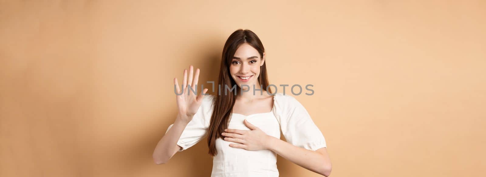 Cute smiling girl make promise, put hand on heart and tell truth, being honest, swearing to you, standing on beige background.