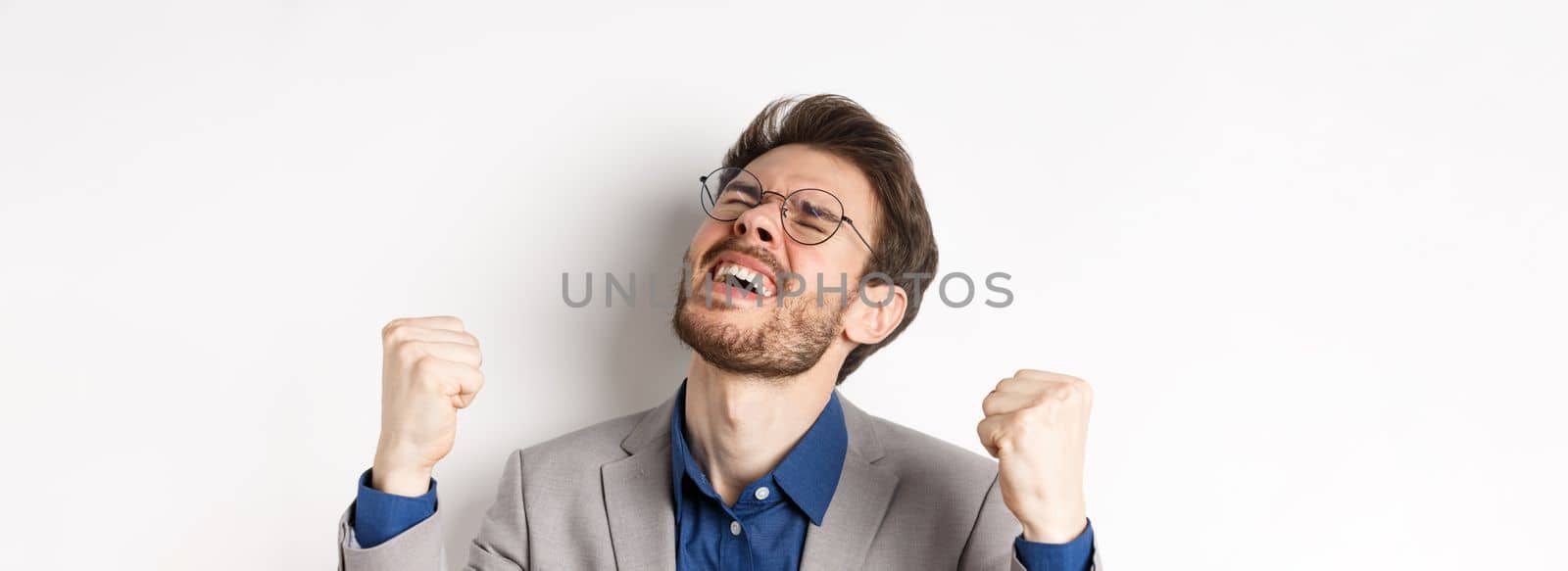 Excited businessman in glasses and suit shouting yes with pleasure and relieved face, shaking fists up, triumphing, winning bet, standing on white background.