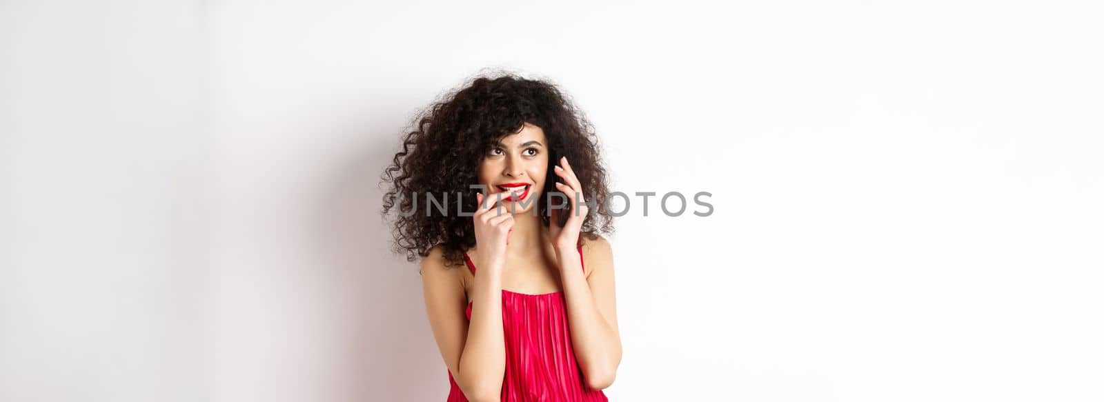 Silly fashionable woman in red dress and lips, biting fingernail duing phone call, thinking on mobile conversation, standing on white background.