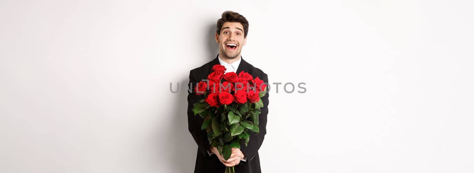 Image of handsome boyfriend in black suit, holding bouquet of red roses and smiling, being on a date, standing over white background by Benzoix