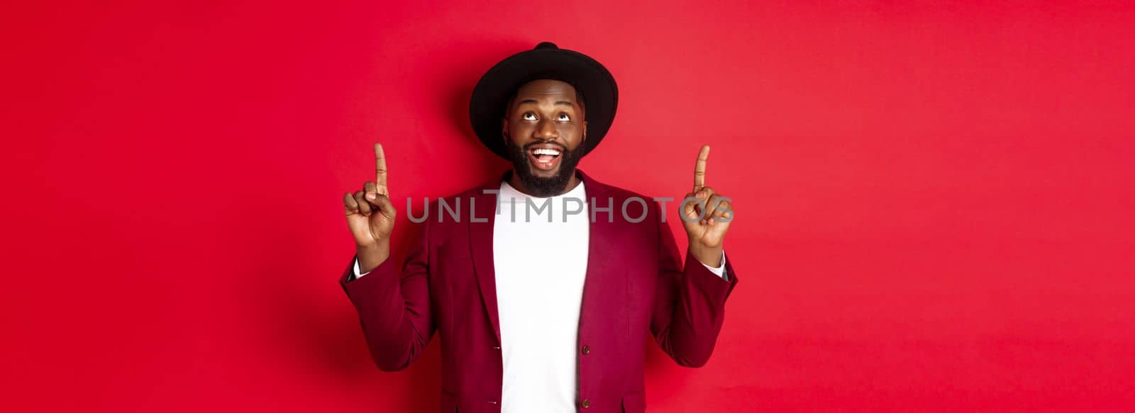 Winter holidays and shopping concept. Cheerful Black man looking and pointing fingers up, checking out xmas promo, standing over red background.