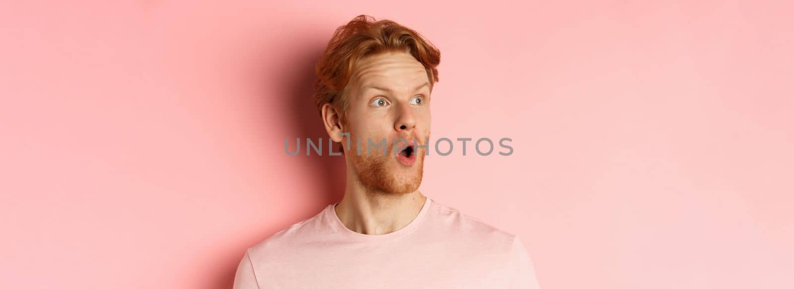 Close up of shocked redhead man with beard, saying wow, looking left with amazed face, standing over pink background.