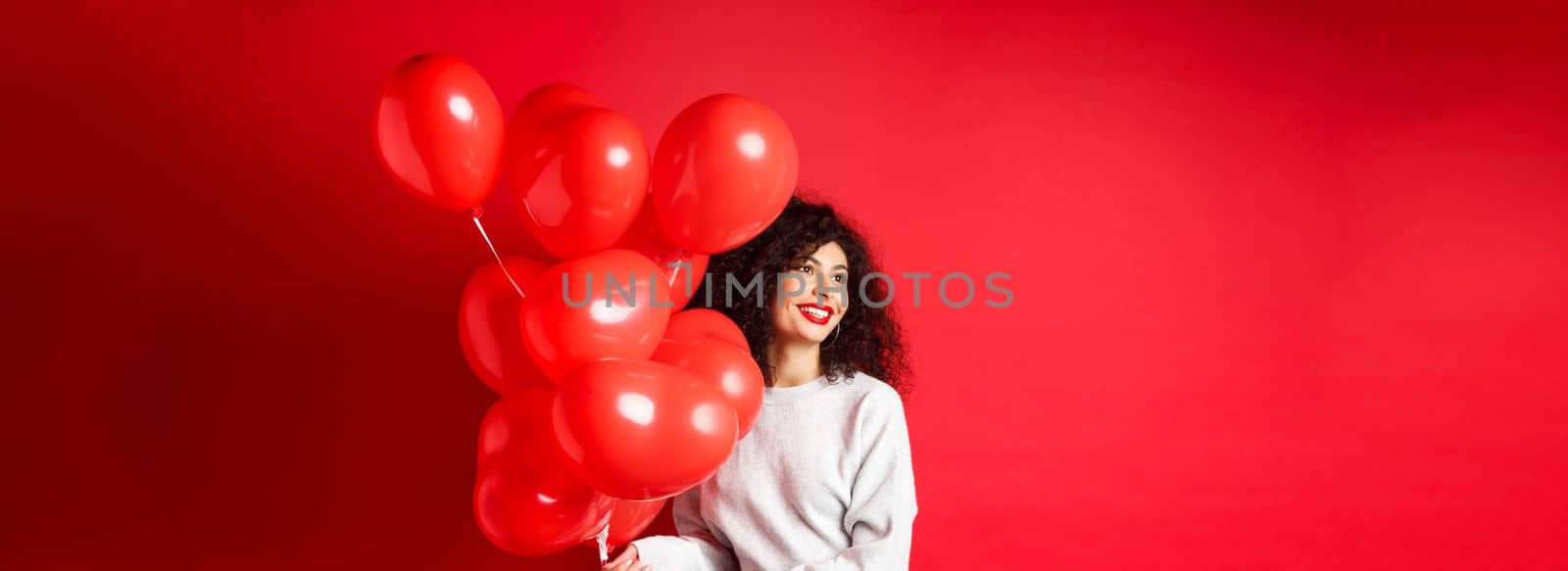 Holidays and celebration. Happy woman posing with party balloons on red background, looking aside at empty space.