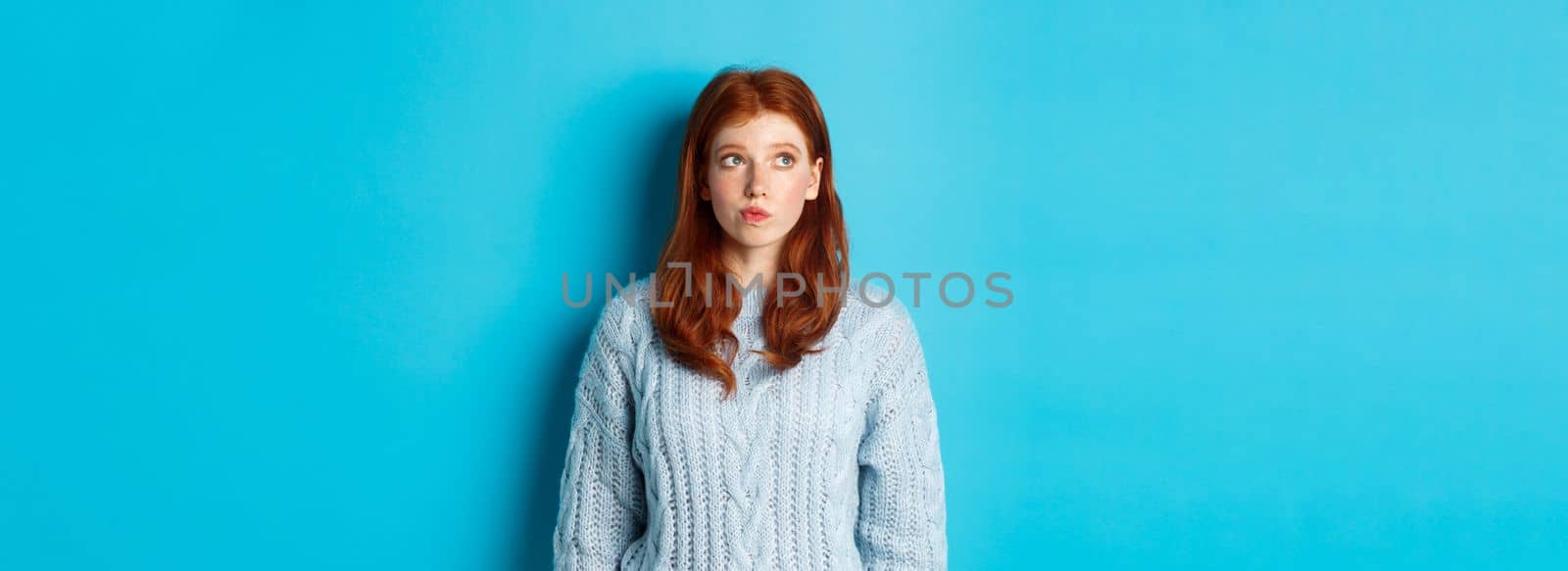 Dreamy redhead girl thinking or making decision, looking at upper left corner logo, standing against blue background.
