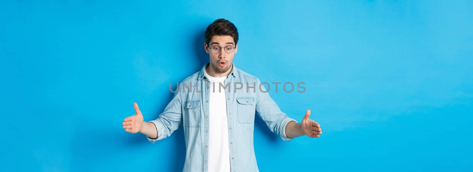 Excited handsome man showing big size object and looking amazed, standing over blue background by Benzoix