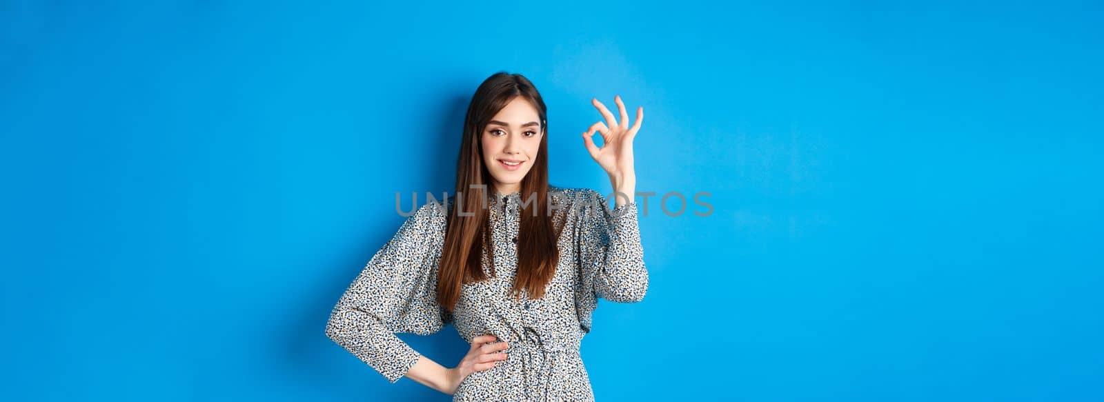Confident young woman in dress showing okay sign and smiling, no problem gesture, assure everything good, standing on blue background.