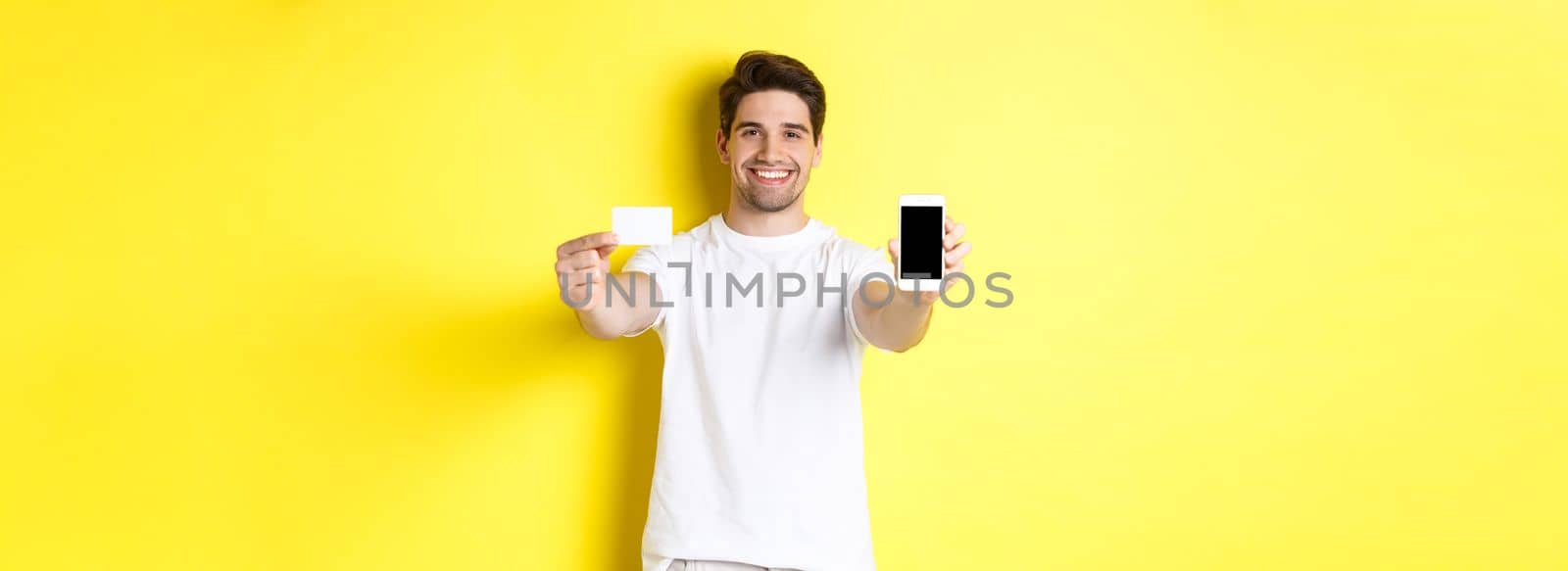 Handsome caucasian male model showing smartphone screen and credit card, concept of mobile banking and online shopping, yellow background.