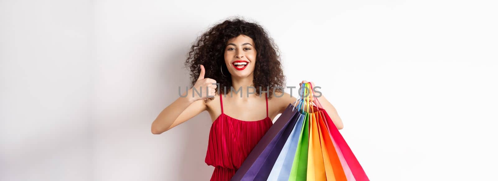 Fashionable woman in red dress going shopping, holding bags and showing thumbs up, recommend store, standing on white background.
