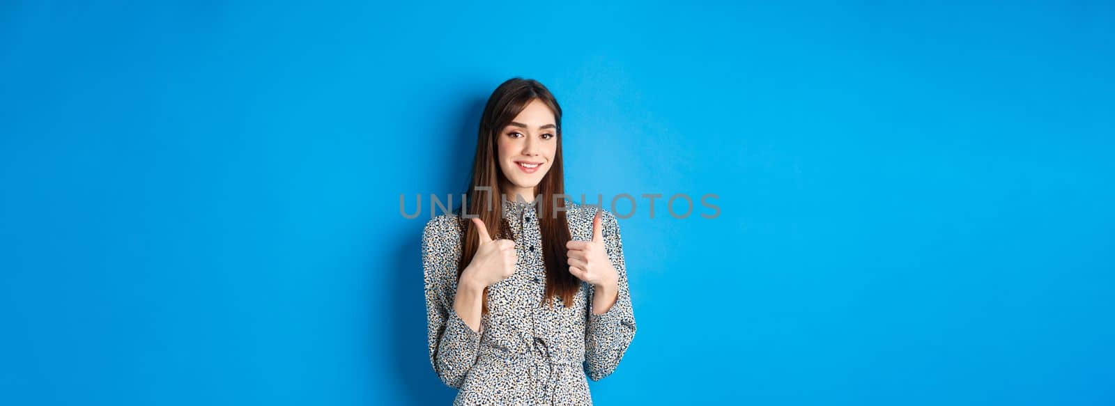 Cheerful smiling woman in dress, showing thumb up and look satisfied, approve and like good thing, recommending product, standing on blue background by Benzoix