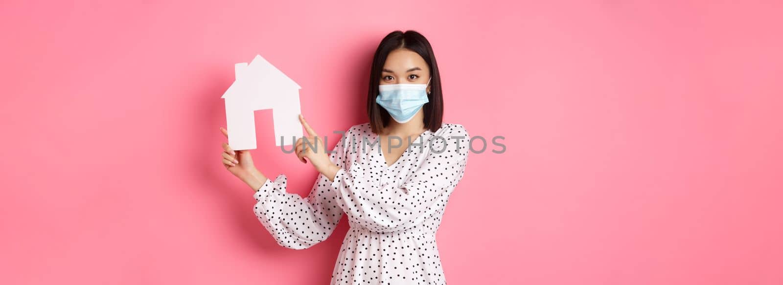 Covid-19, real estate and lifestyle concept. Cute asian woman in face mask selling houses, showing model of home and looking at camera, standing over pink background.