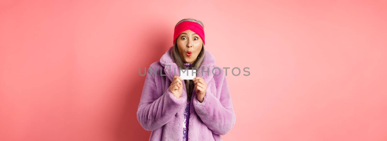 Shopping and fashion concept. Image of amazed asian senior woman checking out promo offer, holding plastic credit card and stare impressed at camera, pink background.