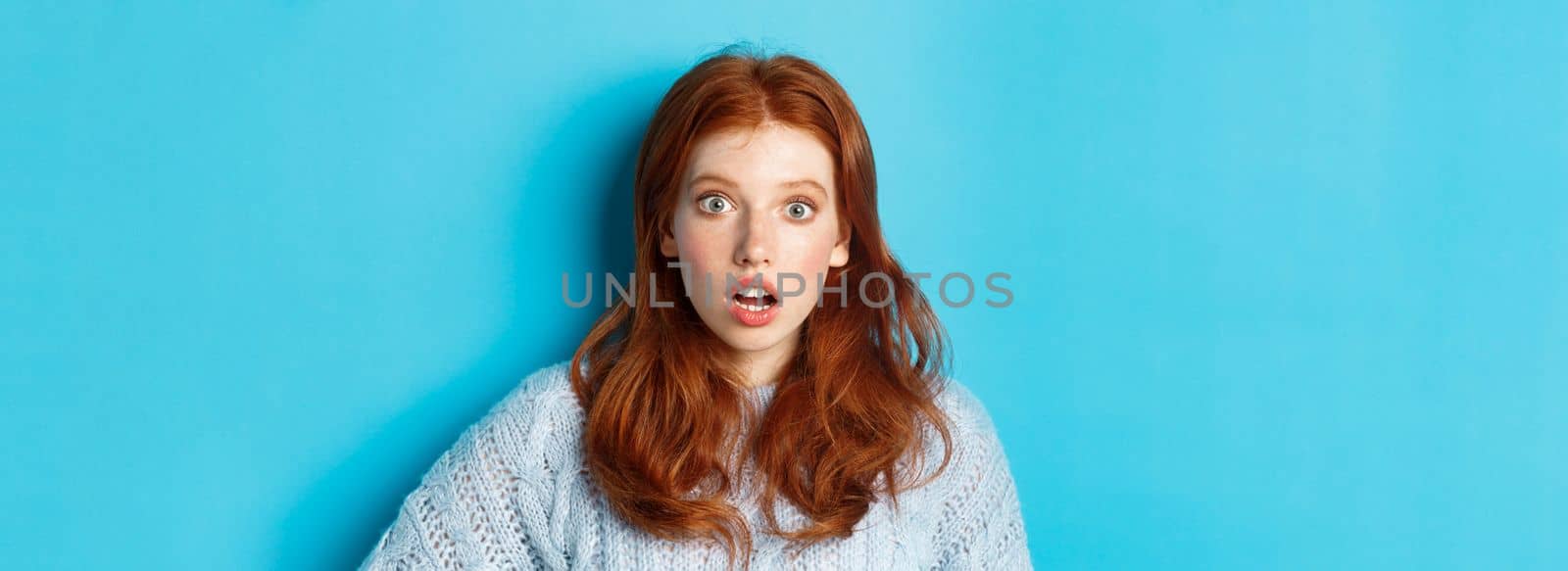 Close-up of startled redhead girl stare at camera speechless, open mouth in awe, standing over blue background.