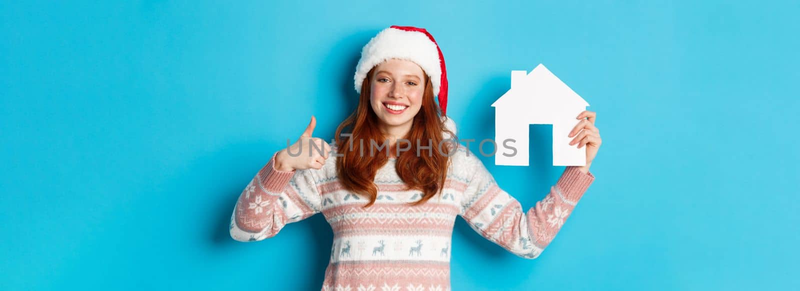 Holiday promos and real estate concept. Satisfied female model with red wavy hair, wearing santa hat and sweater, showing paper house model and thumbs-up, blue background.