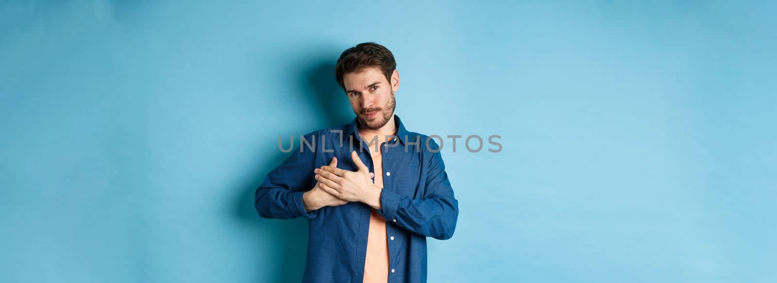 Romantic man holding hands on heart and smiling, thanking you, standing grateful on blue background.