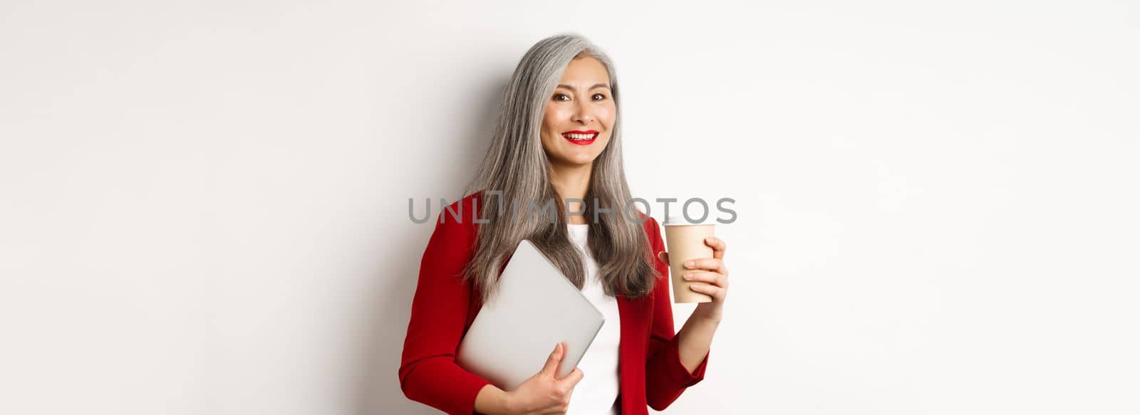 Business. Successful asian businesswoman with grey hair, wearing red blazer, drinking coffee and standing with laptop in hand, white background.