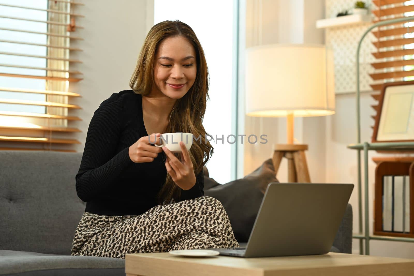 Pleasant beautiful woman in casual clothes drinking coffee and using laptop in living room.