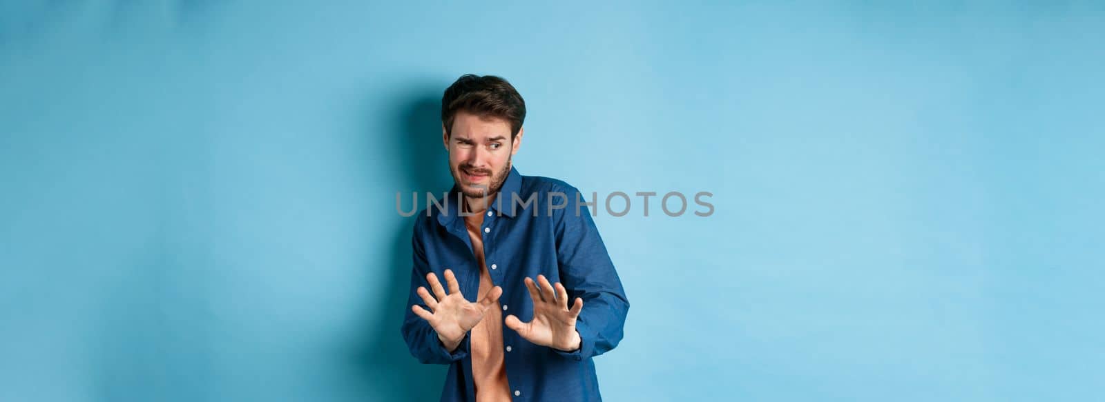 Man cringe and stay away from something nasty, looking aside and stretch out hands to reject bad deal, standing on blue background.