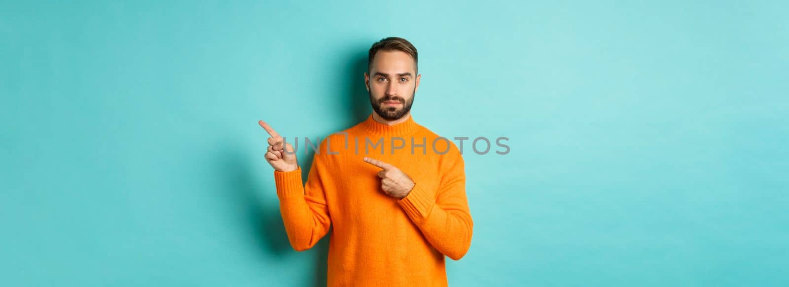 Handsome and serious bearded man in orange sweater pointing right, showing advertisement or logo, standing over turquoise background.