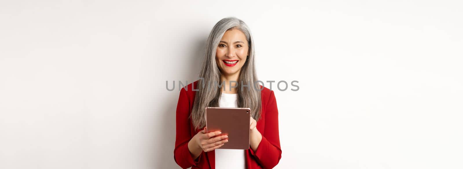 Business. Senior female entrepreneur working on digital tablet and smiling happy at camera, wearing red blazer and makeup, standing over white background.