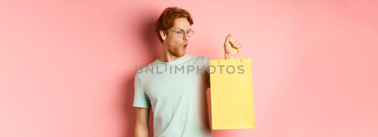 Handsome young man buying presents, holding shopping bag and looking amused, standing over pink background.