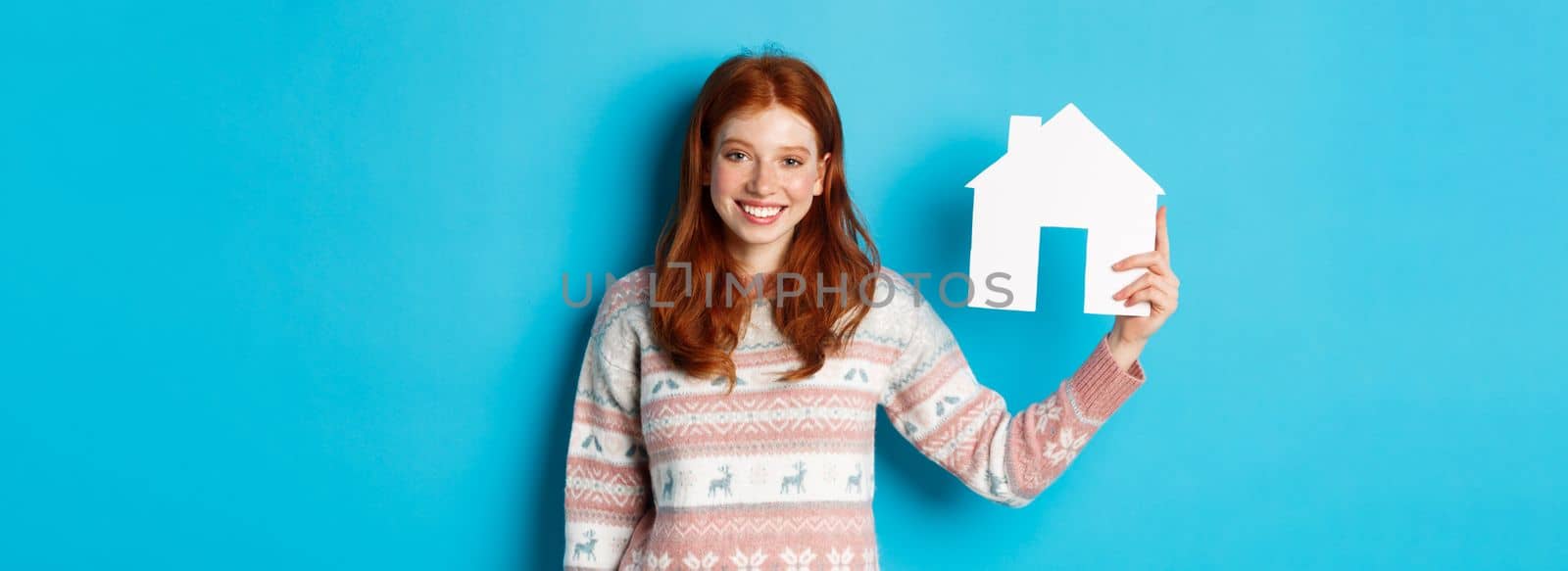 Real estate concept. Young smiling woman with red hair showing paper house model, standing over blue background.