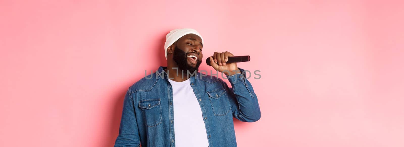 Handsome Black man in beanie and denim shirt singing karaoke, holding microphone, standing over pink background.