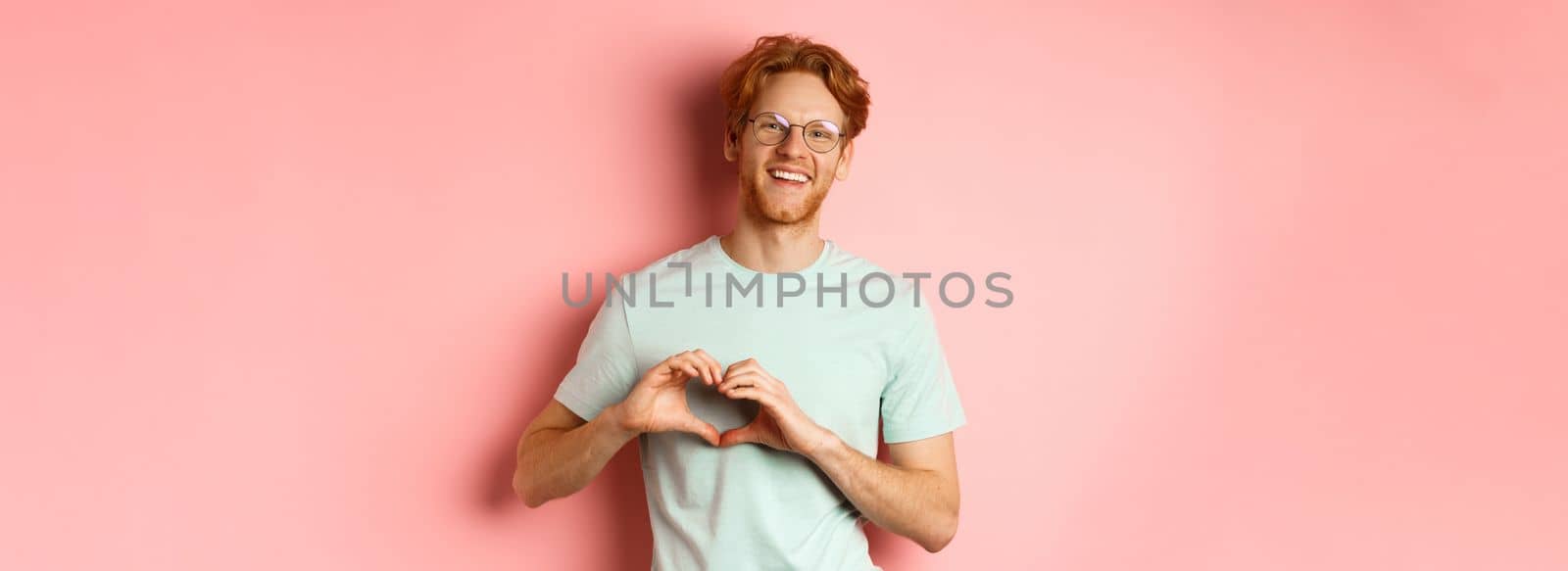 Valentines day concept. Handsome redhead man in glasses, showing heart sign and say I love you, standing over pink background.