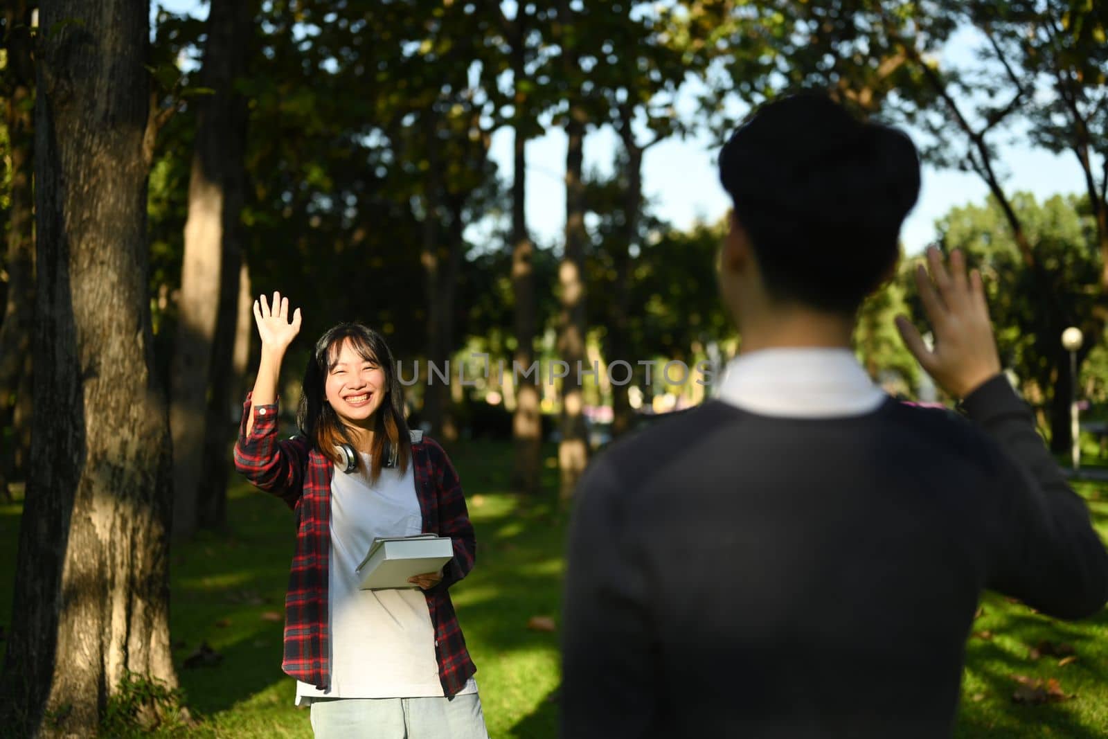 Cheerful asian student woman walking in public park and greeting friend with hand raised up.