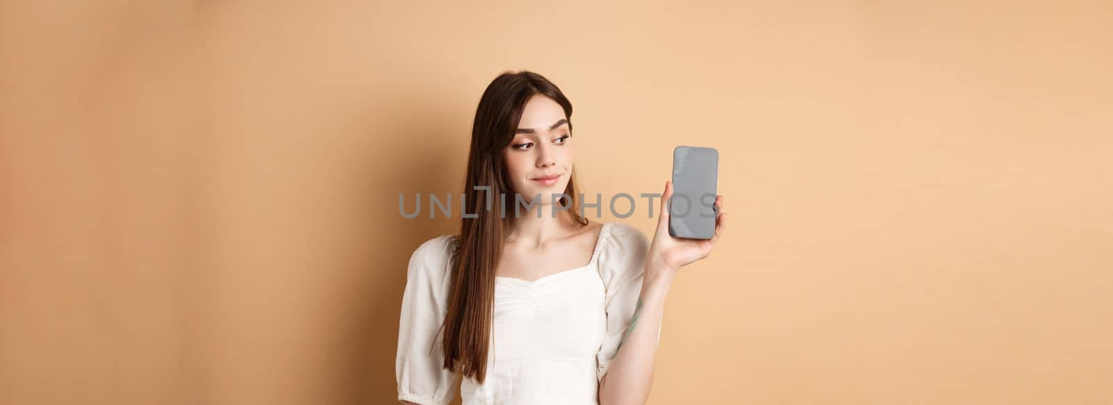 Attractive young woman showing empty smartphone screen, smiling and looking aside, standing on beige background.