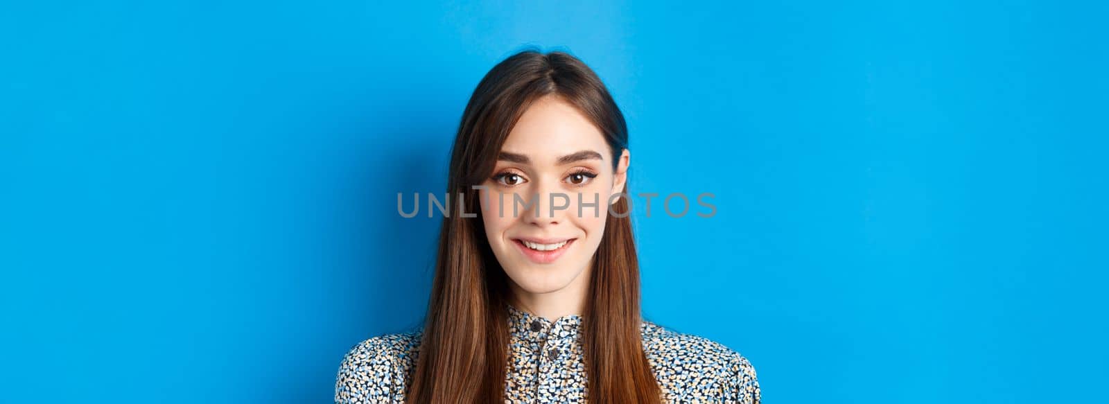 Close-up portrait of attractive natural girl with long hair and makeup, smiling happy and friendly at camera, blue background.