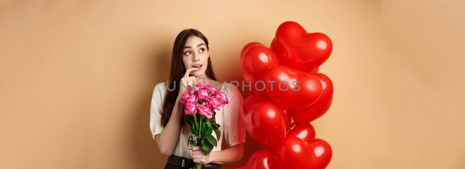 Dreamy young woman holding bouquet of roses and thinking about secret admirer on Valentines day, looking at upper left corner and biting finger, standing near red romantic balloons.
