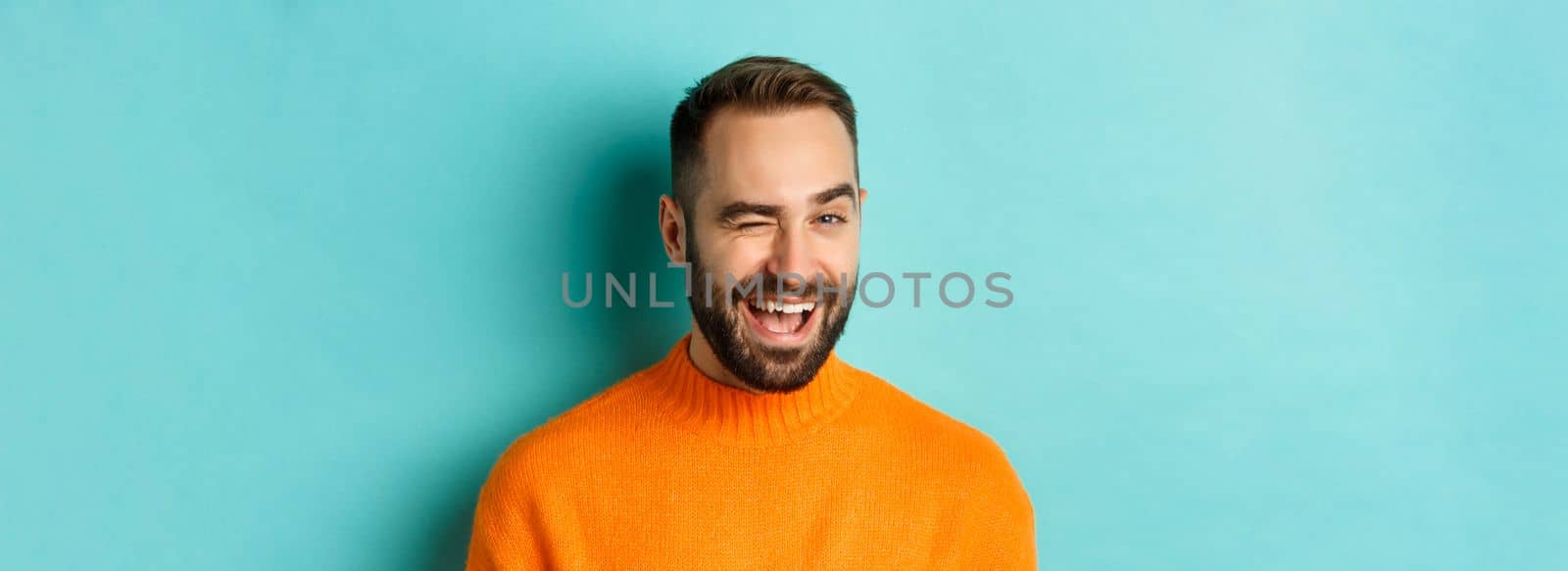 Handsome confident man winking at camera, smiling sassy, standing in orange sweater against blue background.