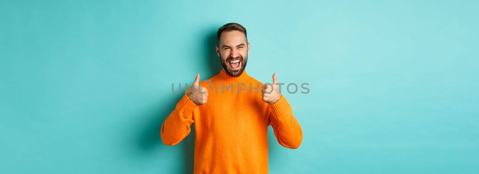 Super good. Satisfied happy man showing thumbs up, agree with you, praise excellent work, looking pleased, standing in orange sweater against light blue background by Benzoix