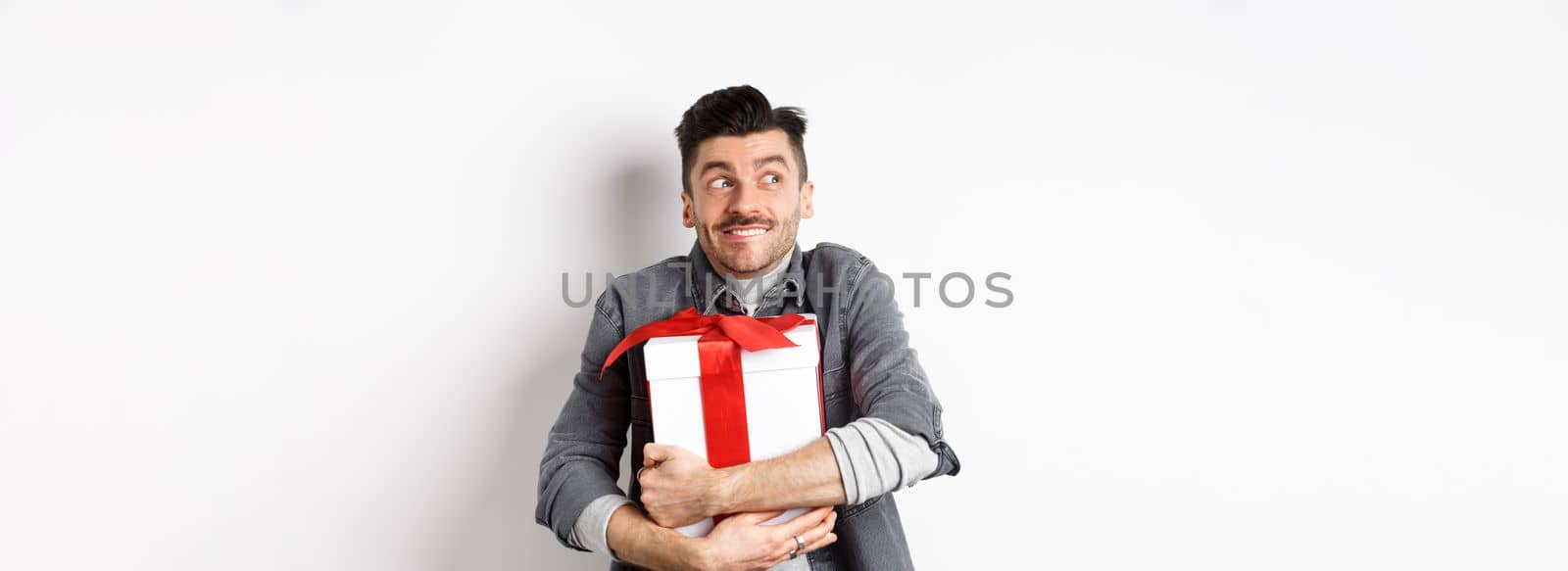 Happy boyfriend hugging gift from lover, looking aside at empty space and smiling excited, making romantic present on valentines day, standing on white background.