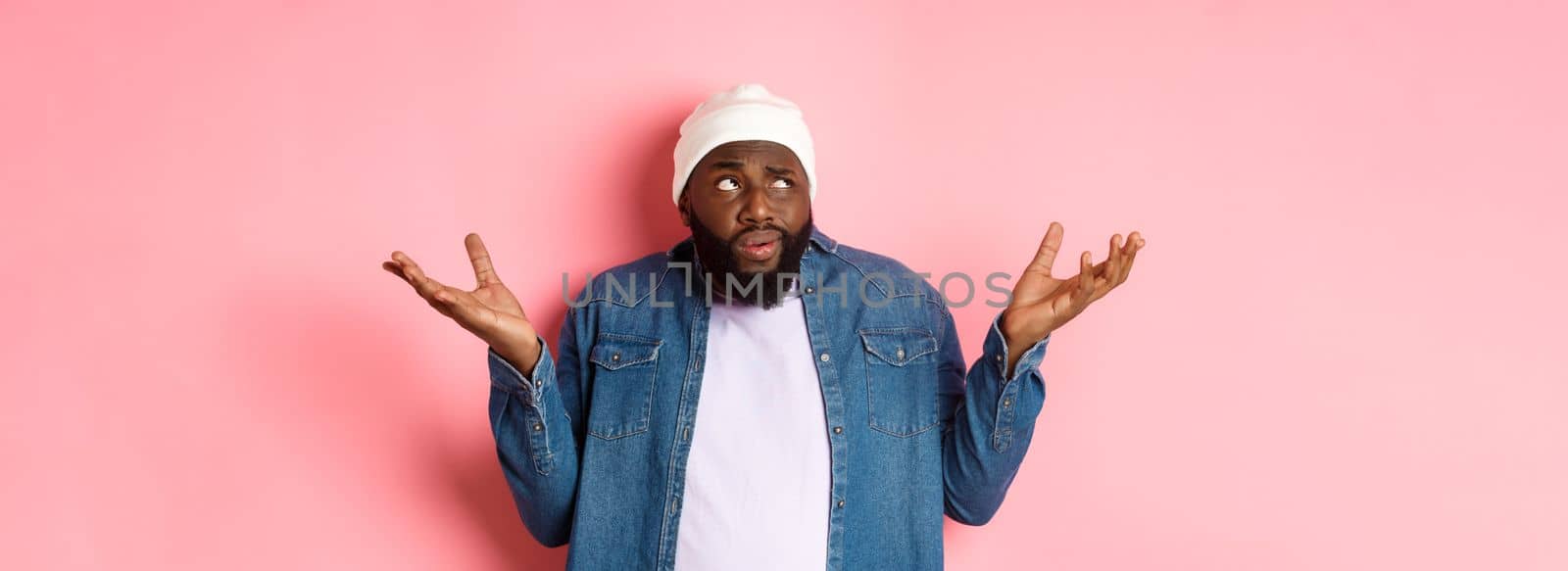 Confused african-american man in beanie looking at upper left corner doubtful, shrugging uncertain, standing over pink background.