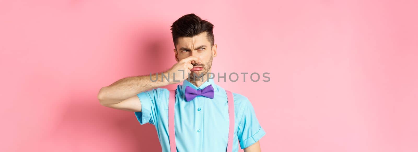 Disgusted young man in bow-tie and suspenders shut his nose from awful smell, looking at smelly thing, standing over pink background.