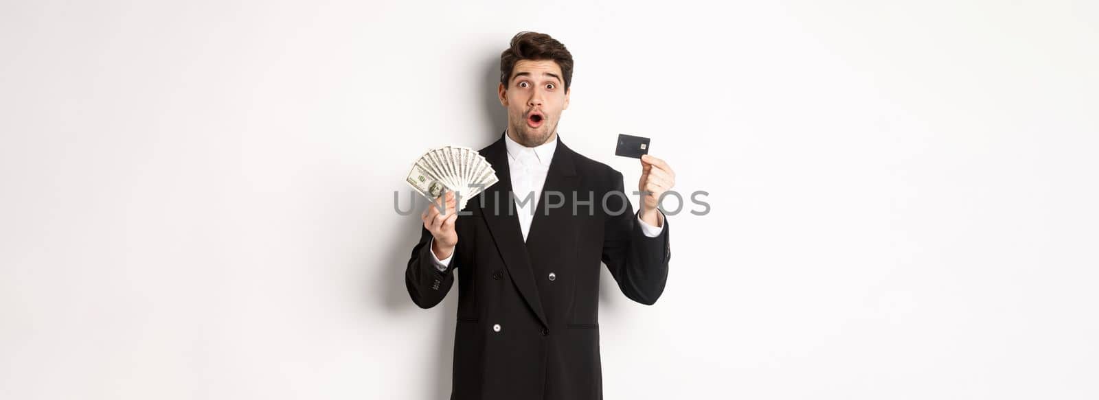 Portrait of surprised handsome man i suit, showing credit card with money, standing against white background by Benzoix