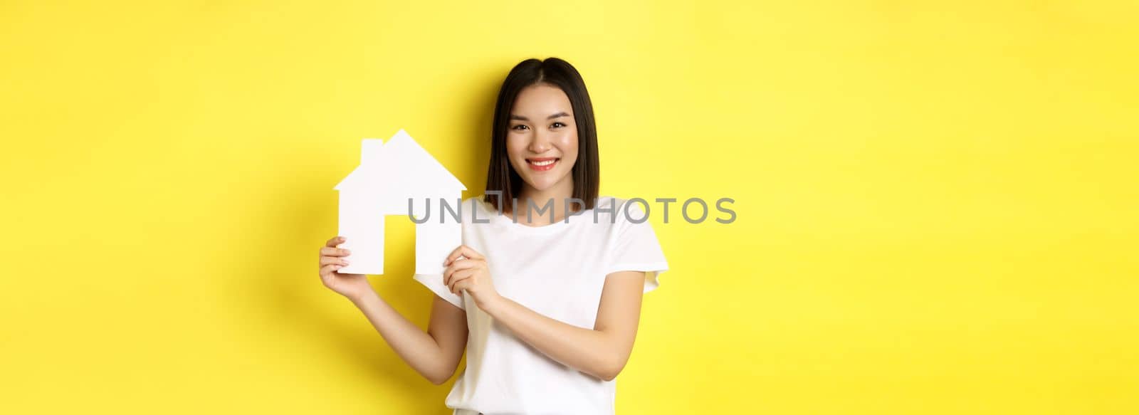 Real estate concept. Smiling beautiful woman showing paper house cutout and looking at camera, buying property, standing over yellow background by Benzoix